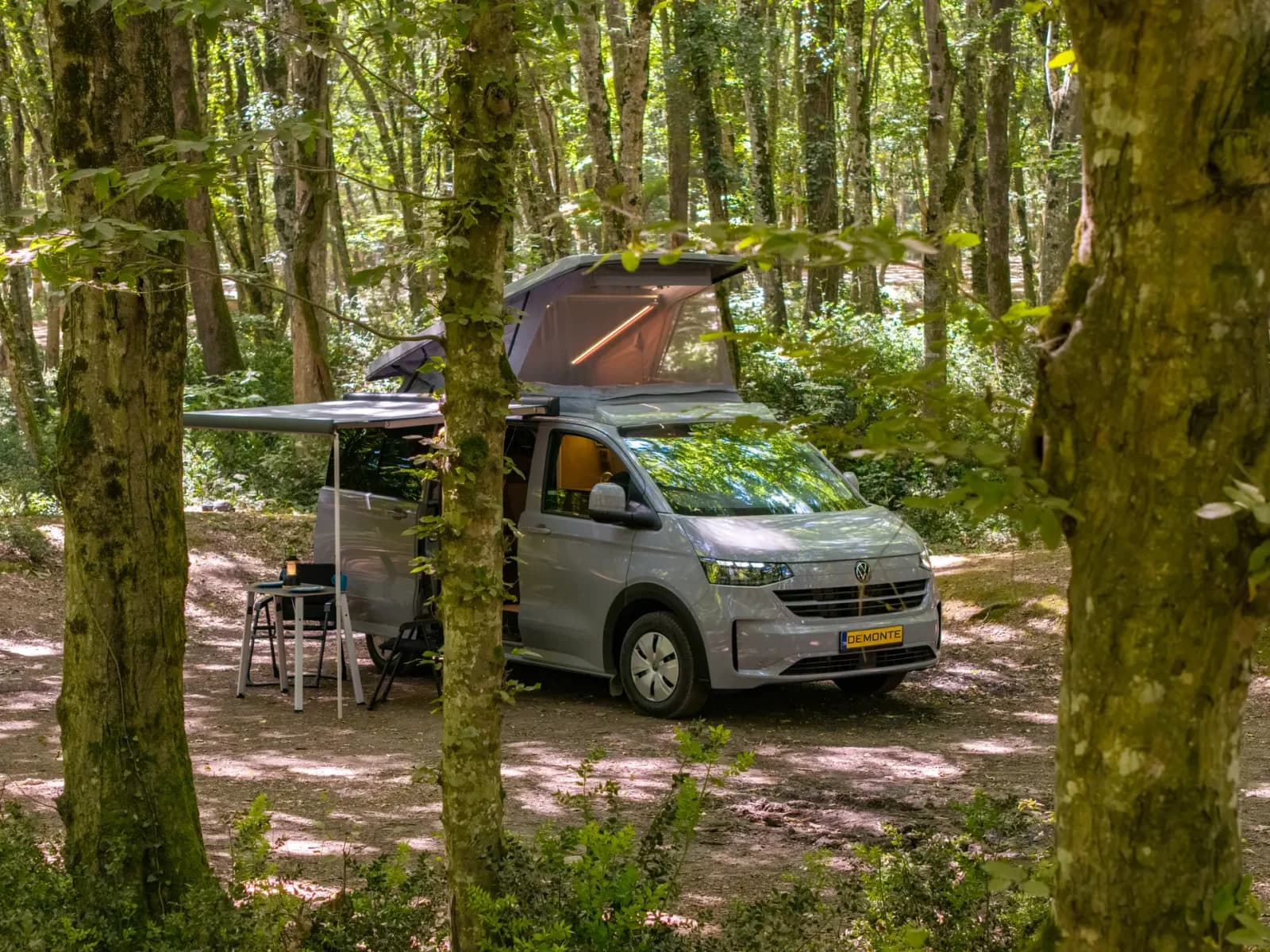 Demonte-Camper zwischen den Bäumen, das Aufstelldach vollständig aufgestellt und das Camp unter der Markise aufgebaut