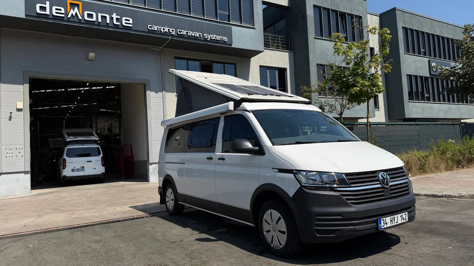 Weißer VW Transporter Efes vor dem Demonte-Caravan-Werk mit SCA-Aufstelldach und Solarmodul