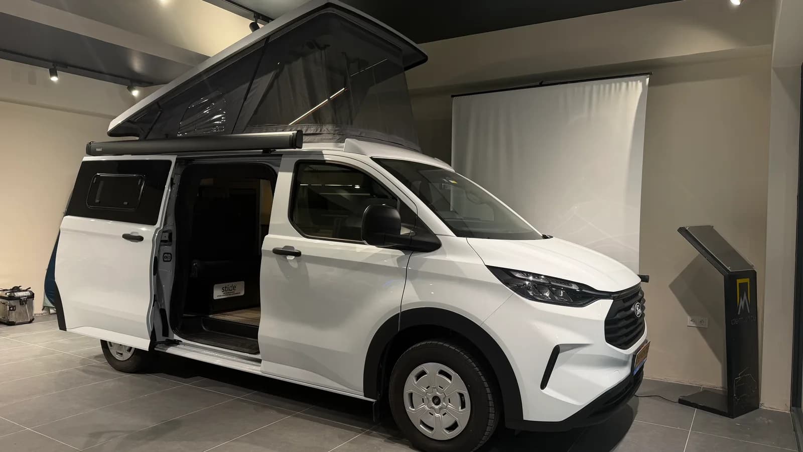 Weißer Ford Transit Custom Assos im Showroom mit ausgestelltem SCA-Aufstelldach und offener Schiebetür